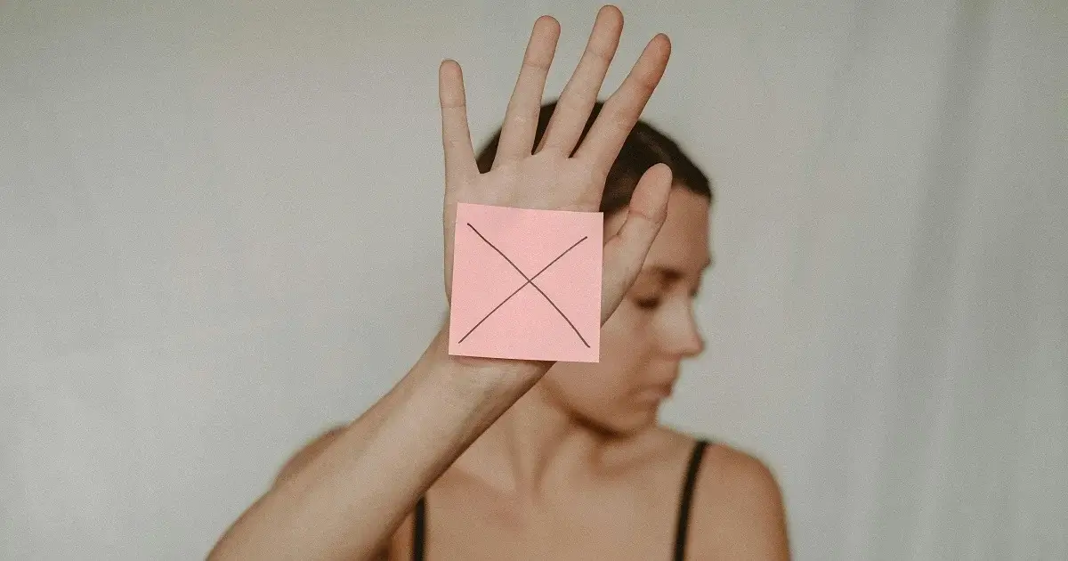 A girl holding up her hand with a sticky note with an x on, symbolizing `no`